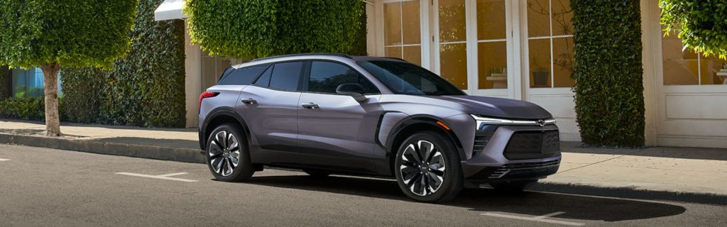 2025 Chevrolet Blazer EV parked on a city street in front of a building with greenery, showcasing its sleek aerodynamic design, bold front fascia, and modern alloy wheels.